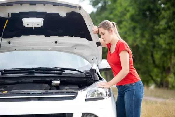 Sad young woman leaning on open hood of her broken car at countryside road — Stock Photo, Image