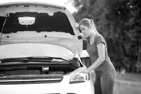 Black and white portrait of young upset woman leaning on open hood of her broken car — Stock Photo, Image