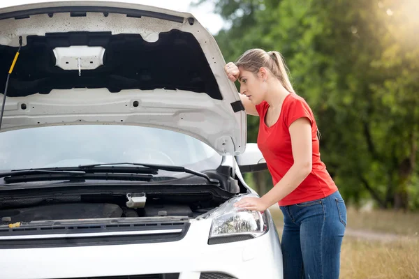 Portrait of sad young woman leaning on open car hood and looking at broken engine — Stock Photo, Image