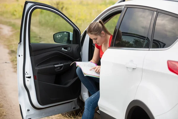 Mujer joven sentada en el coche en la carretera rural mirando mapas de carreteras — Foto de Stock