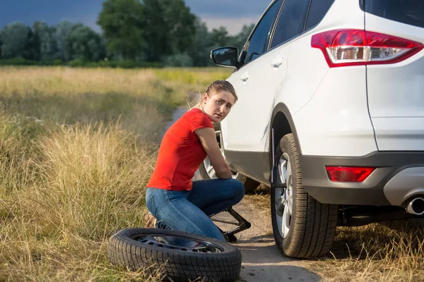 Jeune femme bouleversée soulevant sa voiture cassée avec cric pour changer de pneu plat sur la route de campagne — Photo