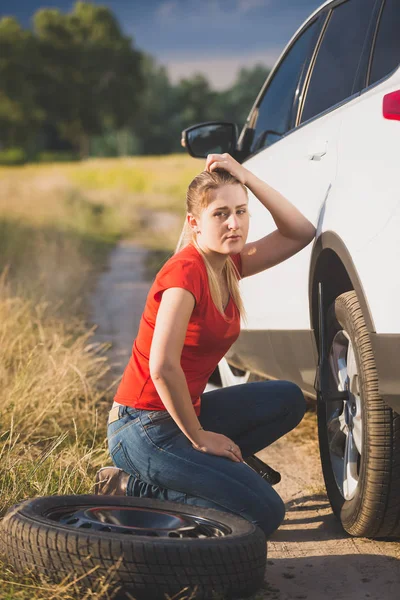 Foto tonificada de una triste joven tratando de cambiar el neumático pinchado del coche en la carretera desierta en el campo —  Fotos de Stock