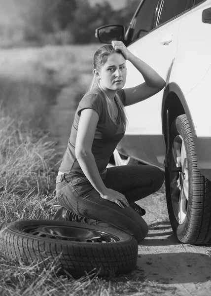 Black and white image of young woman sitting at car with flat tire and looking in camera — Stock Photo, Image