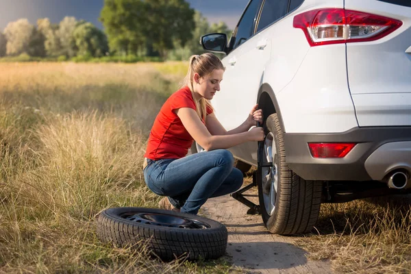 Mujer joven cambiando rueda de repuesto del coche en la carretera desierta en el campo — Foto de Stock