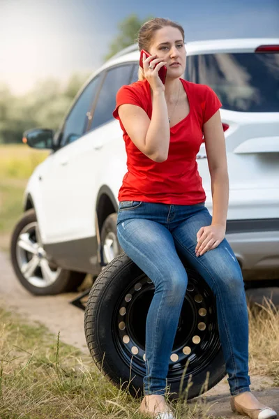Young woman sitting on car spare wheel at her broken transport and calling service — Stock Photo, Image