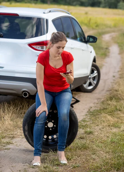 Mujer joven con coche roto sittign en la rueda de repuesto y llamando al servicio automático para obtener ayuda —  Fotos de Stock