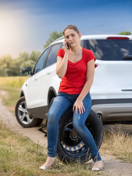 Smutny młoda kobieta siedzi obok samochodu z złamane koło auto zamówimy — Zdjęcie stockowe