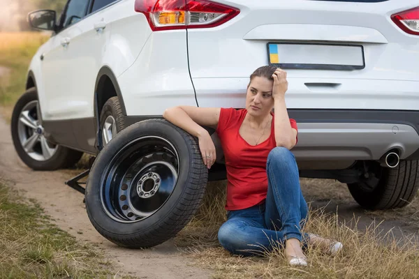 Giovane donna seduta accanto all'auto con pneumatico piatto in campo e in attesa di aiuto — Foto Stock
