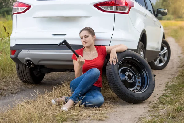 Young woman sitting at broken car in field and holding car jack — Stock Photo, Image