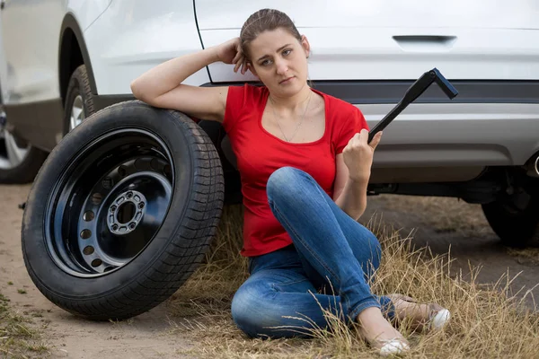 Retrato de una joven sentada en el caar roto en el campo y sosteniendo la llave inglesa de la rueda del coche —  Fotos de Stock