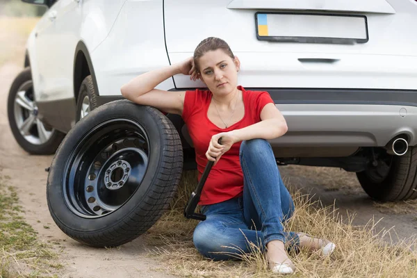 Jovem mulher chateada sentada no chão ao lado de carro quebrado na estrada deserta e esperando por ajuda — Fotografia de Stock