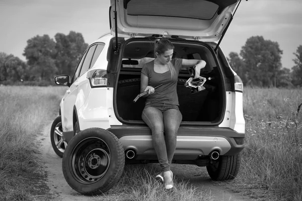 Imagem em preto e branco da jovem mulher sentada em SUV tronco aberto e olhando para a chave de rodas e jack — Fotografia de Stock