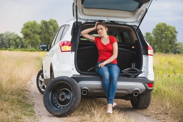 Mujer joven sentada en el maletero abierto y sosteniendo herramientas para cambiar neumáticos pinchados —  Fotos de Stock