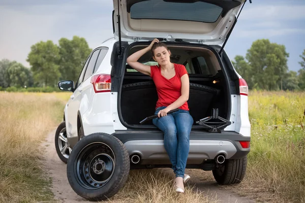 Mujer joven molesta sentada en el maletero del coche abierto y esperando ayuda para cambiar el neumático pinchado —  Fotos de Stock