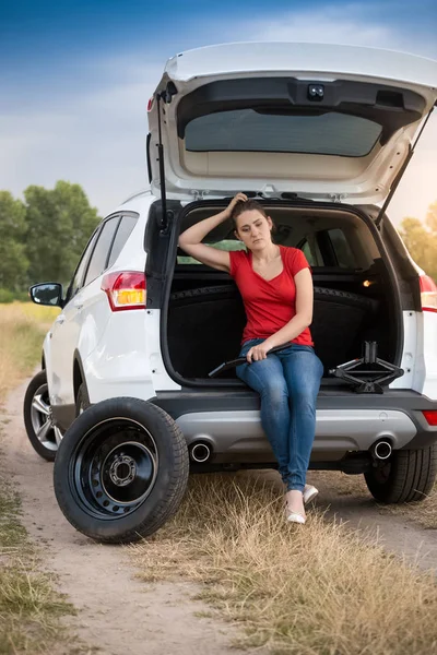 Mujer joven sentada en el maletero del coche abierto esperando que el servicio automático cambie el neumático pinchado — Foto de Stock