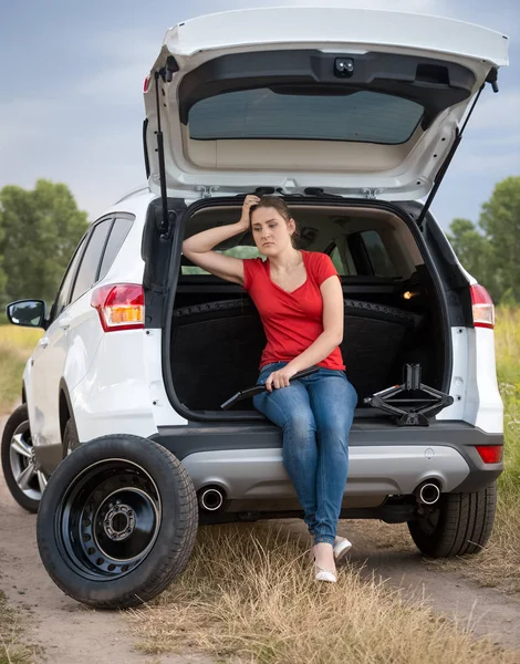 Upset young woman sitting in open trunk of broken car in field — Stock Photo, Image