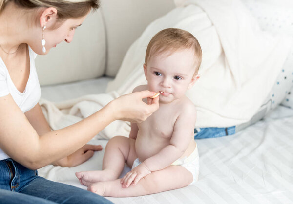 Cute baby boy sitting on bed with mother and eating cookies
