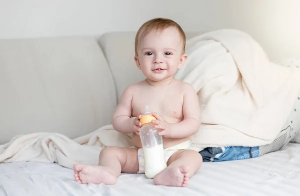 Portrét roztomilý usměvavý chlapeček sedí na posteli a drží láhev mléka — Stock fotografie