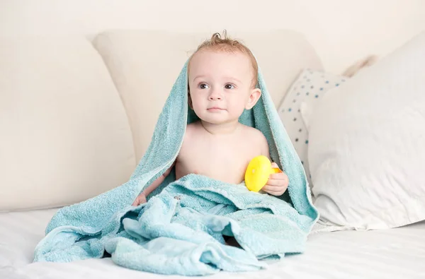 Retrato de menino adorável coberto de toalha azul sentado na cama com pato de borracha amarela — Fotografia de Stock