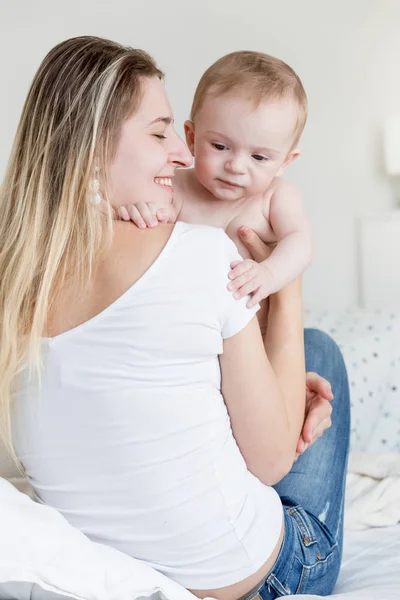 Portriat of happy laughing young owman sitting on bed and holding her 9 months old baby son — Stock Photo, Image