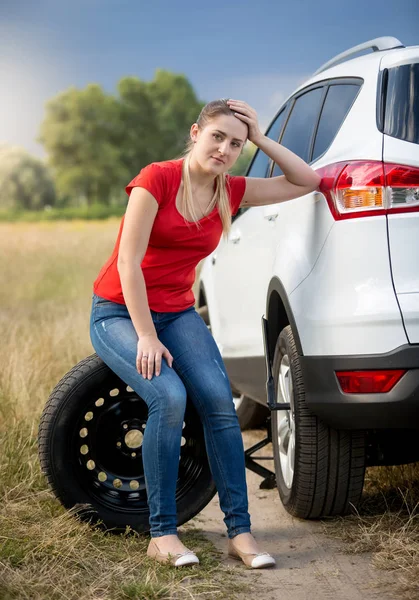 Triste jovem mulher sentado carro roda sobresselente na estrada deserta no campo Imagens Royalty-Free