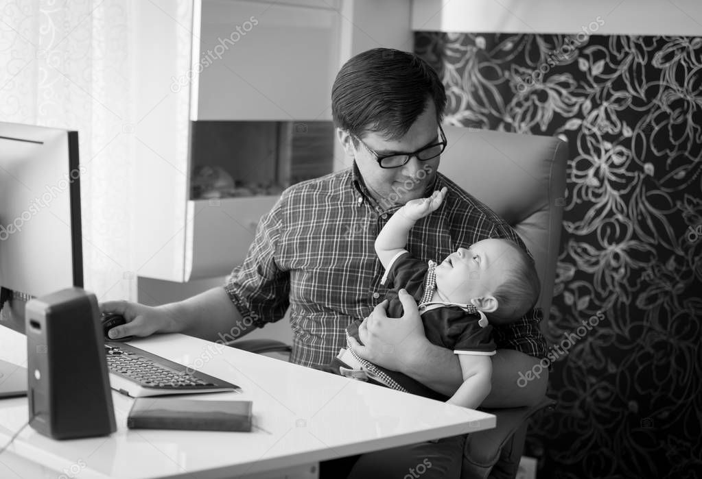 Black and white image of smiling young man working in home office and looking after his baby son