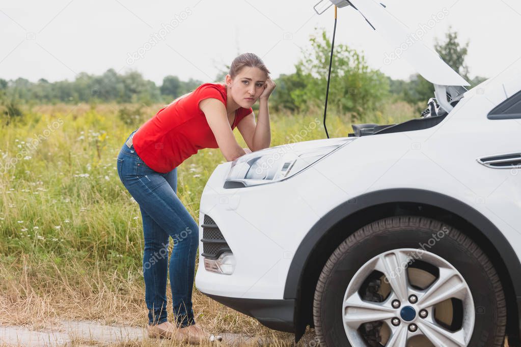 Portrait of sad young woman leaning on engine of broken car in field