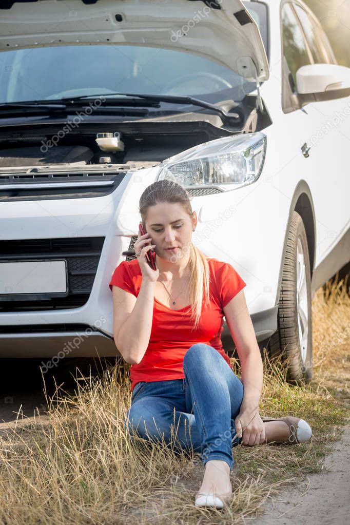 Upset young woman sitting on ground next to broken car calling car service for help
