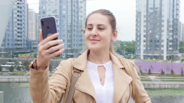 4k-Video der schönen lächelnden jungen Frau, die auf dem Dach steht und ein Selfie mit dem Smartphone vor Wolkenkratzern und der Skyline der Stadt macht — Stockvideo