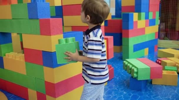 4k-Video von kleinem Jungen, der einen hohen Turm aus großen bunten Blöcken auf einem Spielplatz in einem Einkaufszentrum baut — Stockvideo