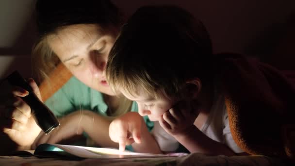 Closeup 4k vídeo de menino deitado na cama com sua mãe à noite e lendo livro com tocha — Vídeo de Stock