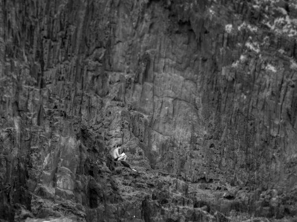 Černobílý obraz mladého osamělého muže sedícího na útesu na břehu moře a čtecí knihy — Stock fotografie