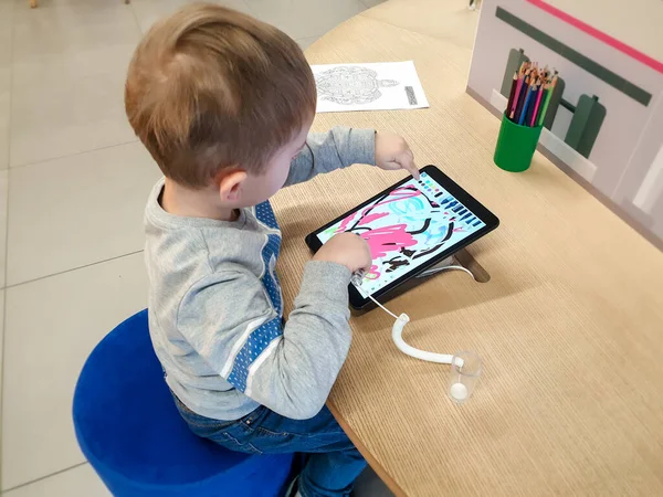Изображение милого 3-летнего мальчика, сидящего за столом и рисующего стилусом на цифровом планшетном компьютере — стоковое фото