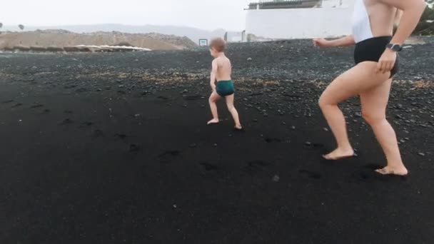 4k видео милого маленького мальчика с красивой молодой матерью работает и играет на черном вулканическом песке пляж океана — стоковое видео