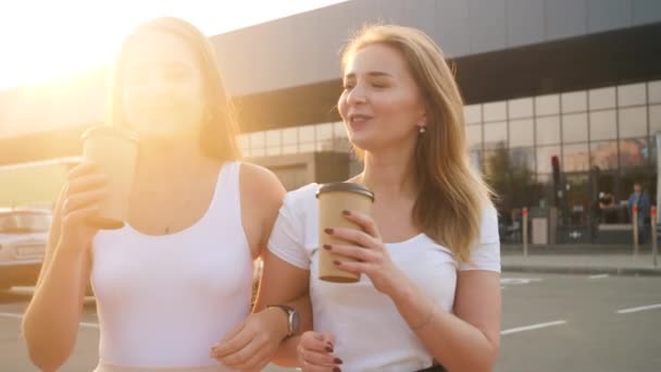 Close up 4k video of two happy girls smiling, talking and drinking coffee while walking on parking lot at shopping mall on the sunset — стоковое видео