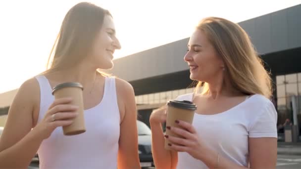 Vidéo 4k de deux amies souriantes et riantes prenant du bon temps et buvant du café tout en marchant dans la rue au coucher du soleil — Video