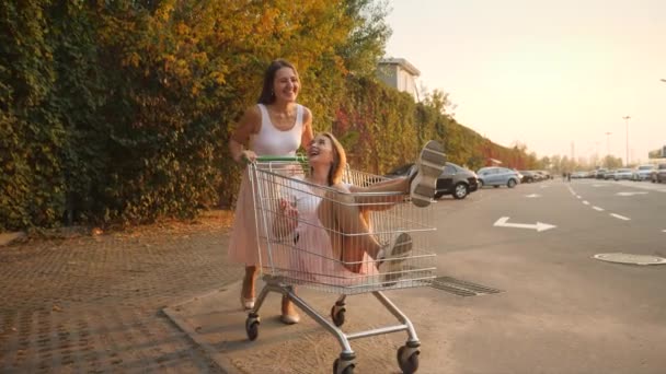 4k-Video von zwei lachenden und lächelnden Freundinnen, die Spaß haben, während sie im Einkaufswagen auf dem Parkplatz eines Einkaufszentrums fahren — Stockvideo