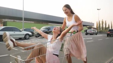 Birbirlerine binen ve otoparkta alışveriş arabasında eğlenen iki genç kızın 4k yavaş çekim videosu.