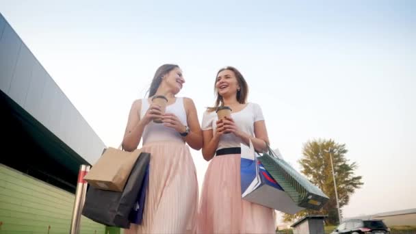 4k χαμηλή άποψη βίντεο από δύο χαρούμενα και γελαστά κορίτσια που περπατούν στο δρόμο της πόλης με πολλές τσάντες ψώνια και πίνοντας καφέ — Αρχείο Βίντεο