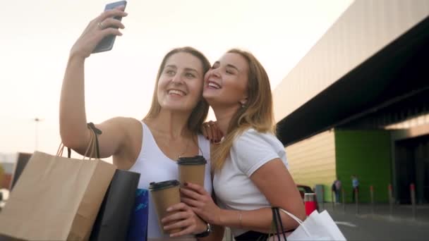 4k крупным планом видео двух счастливых улыбающихся девушек, делающих селфи на камеру смартфона после совершения покупок в большом торговом центре — стоковое видео