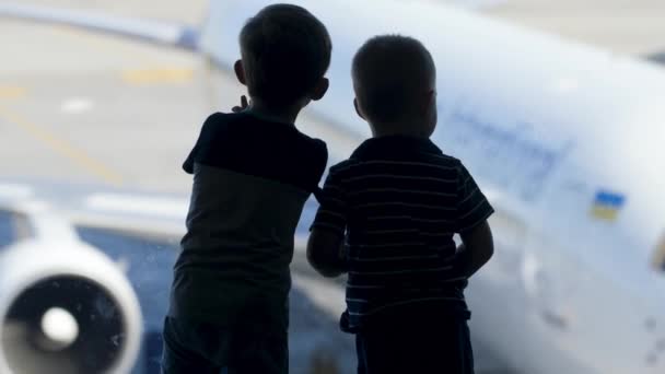 Imágenes de silueta de dos niños pequeños mirando por la ventana grande en la terminal del aeropuerto en el avión esperando a los pasajeros de embarque — Vídeos de Stock