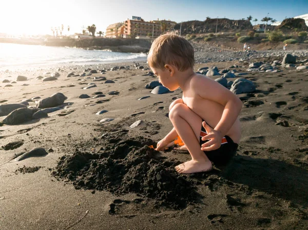 Mały chłopiec siedzi na czarnej plaży wulkanicznej i bawi się piaskiem — Zdjęcie stockowe