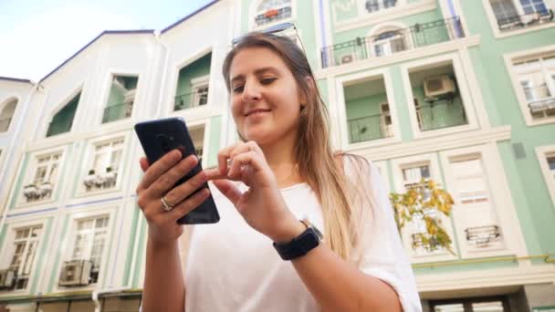 Closeup low angle video of beautiful smiling girl standing on city street and using smartphone — Stockvideo