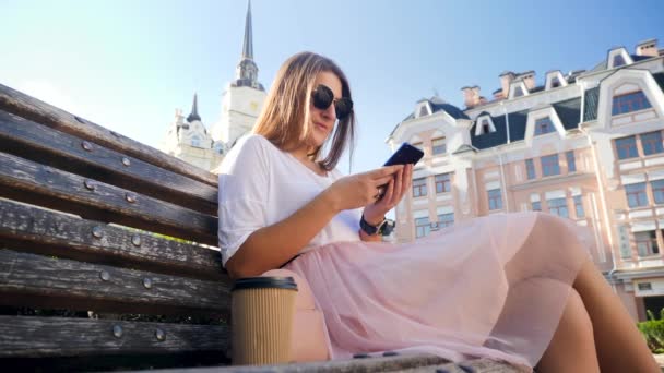 Closeup 4k vídeo de baixo ângulo de mulher jovem digitando mensagem ou navegando na internet no smartphone enquanto sentado no banco no parque e bebendo café — Vídeo de Stock