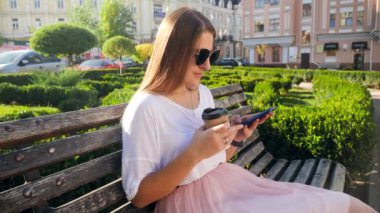 4k slow motion video of beautiful smiling girl drinking coffee and using smartphone while relaxing on bench at city park