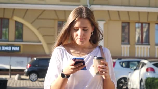 4k video of young beautiful woman with long hair walking on street, using smartphone and drinking coffee to go from paper cup — Stockvideo