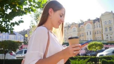 Closeup 4k video of beautiful smiling girl drinking coffee and typing message on smartphone while walking in city on warm sunny day