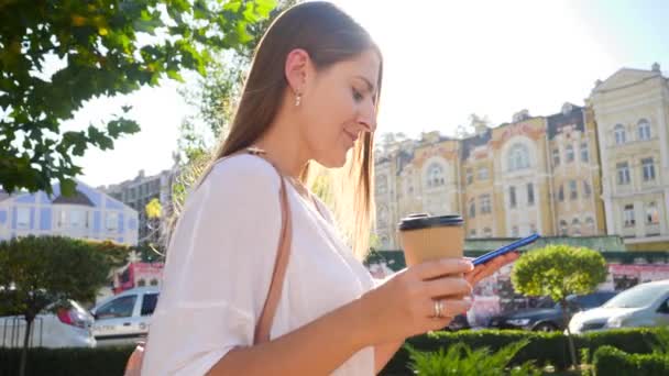 Closeup 4k video of beautiful smiling girl drinking coffee and typing message on smartphone while walking in city on warm sunny day — Stockvideo