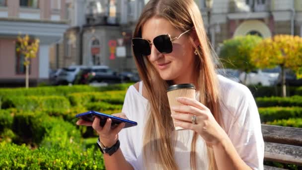 4k video en cámara lenta de la hermosa mujer sonriente en gafas de sol con el pelo largo sentado en el banco en el parque, escribiendo el mensaje en el teléfono inteligente y beber café de la taza de papel — Vídeos de Stock