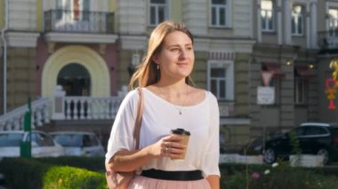 4k slow motion video of happy smiling woman with long hair drinking coffee to go during office break while walking on city street